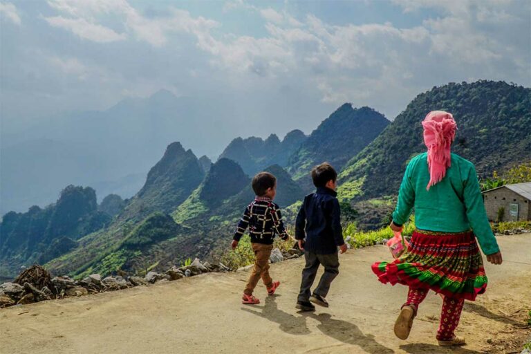 Hmong-vrouw met kinderen op bergpad met uitzicht over groene heuvels en wolken rond de Ha Giang Loop.
