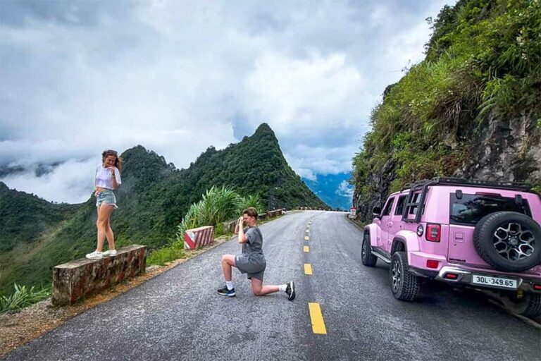 Een reiziger poseert voor een opvallende roze jeep op een bergpas in Ha Giang, met hoge groene pieken en een open bergweg op de achtergrond.