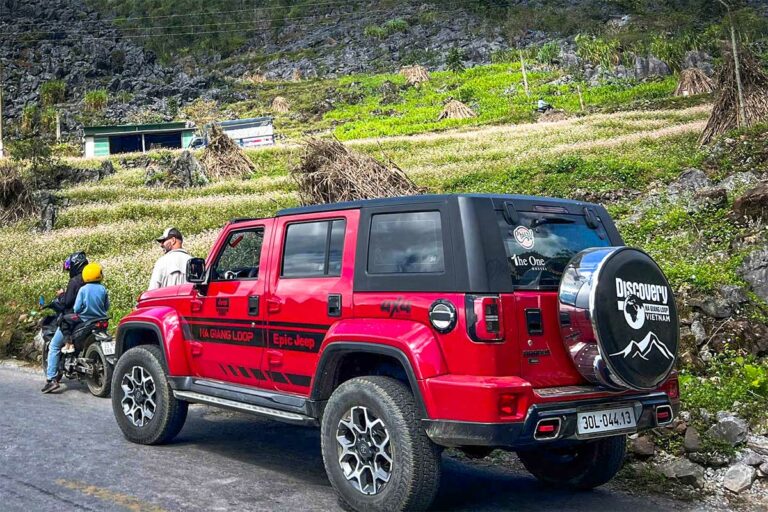 Rode jeep tijdens een Ha Giang Loop tour, geparkeerd langs een weg met uitzicht op rotsachtige heuvels en boekweitvelden.