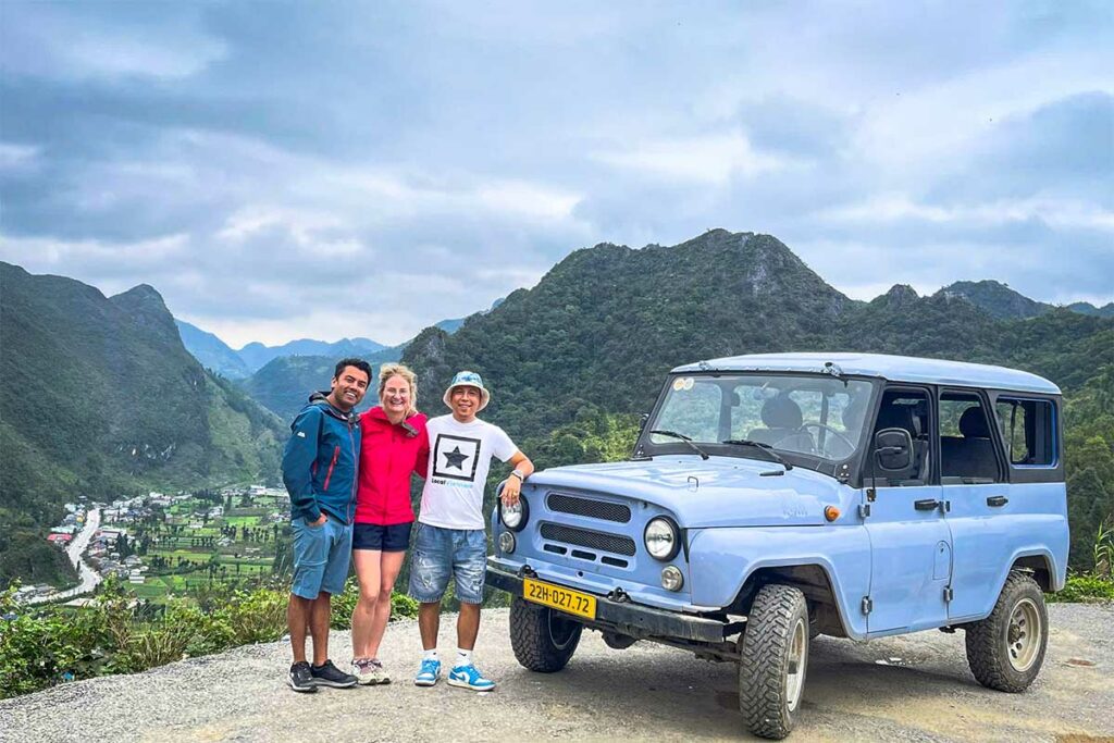 Drie reizigers staan lachend naast een lichtblauwe UAZ-jeep op een uitzichtpunt in Ha Giang, met valleien en karstbergen op de achtergrond.