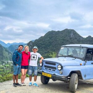 Drie reizigers staan lachend naast een lichtblauwe UAZ-jeep op een uitzichtpunt in Ha Giang, met valleien en karstbergen op de achtergrond.