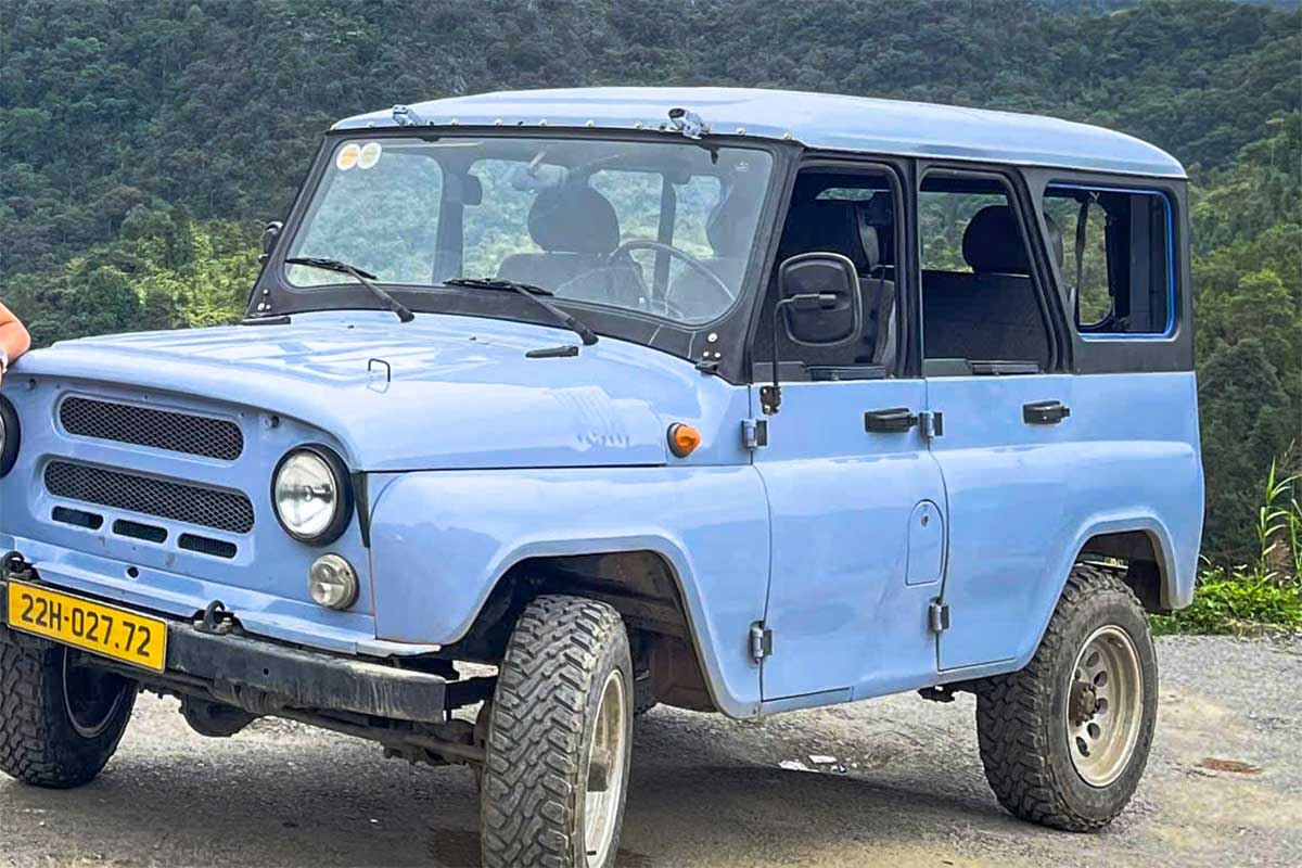 Lichtblauwe UAZ-jeep gebruikt voor Ha Giang Loop tours, geparkeerd met uitzicht op groene bergen.