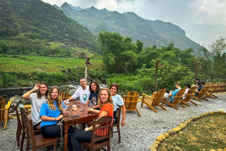 Travelers enjoying drinks at a riverside café in Du Già village surrounded by green mountains.