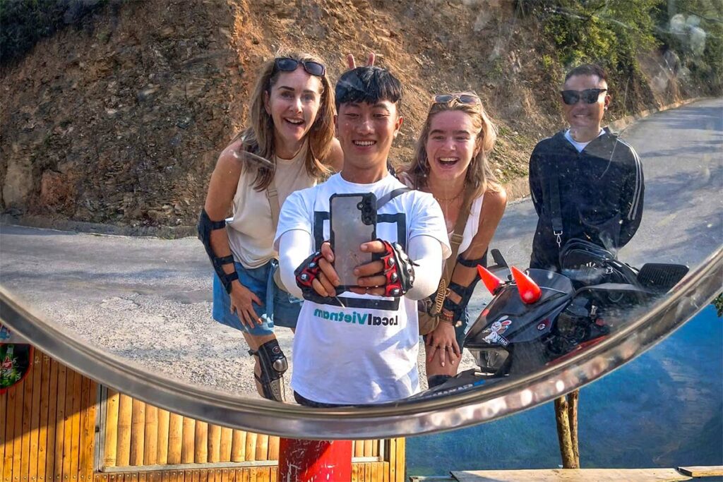 Reizigers en hun Local Vietnam-gids lachen in een spiegelselfie tijdens de Ha Giang Loop Easy Rider tour, met een bochtige bergweg en rotswand in beeld.
