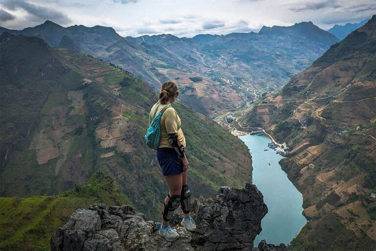 Een reizigster staat op de Ma Pi Leng Sky Walk tijdens de Ha Giang Loop groepstour, uitkijkend over diepe valleien, hoge bergwanden en de kronkelende Nho Que-rivier.
