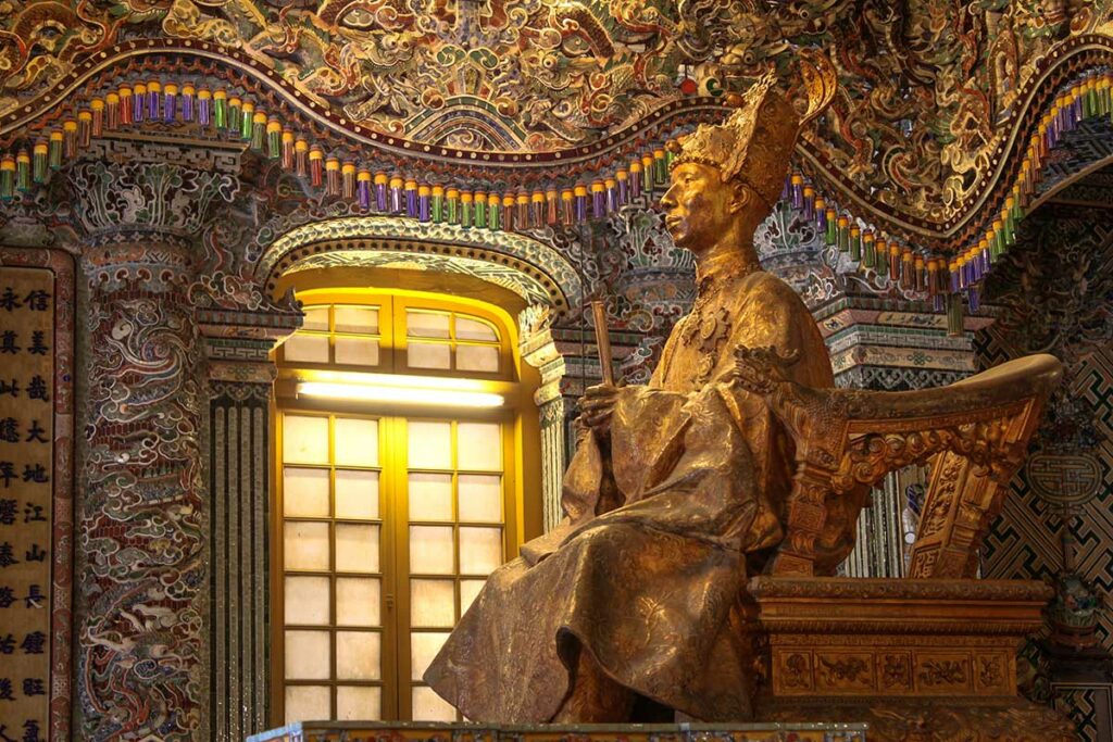 Bronzen zittend beeld van keizer Khai Dinh in Khai Thanh Hall binnen de Tombe van Khai Dinh, omringd door rijk versierde mozaïeken.