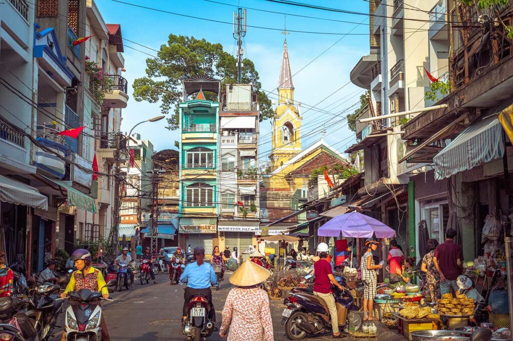 Drukke straat in Chinatown (Cho Lon) in Ho Chi Minh City met scooters, straatverkopers en traditionele gebouwen, met op de achtergrond Cha Tam Church.