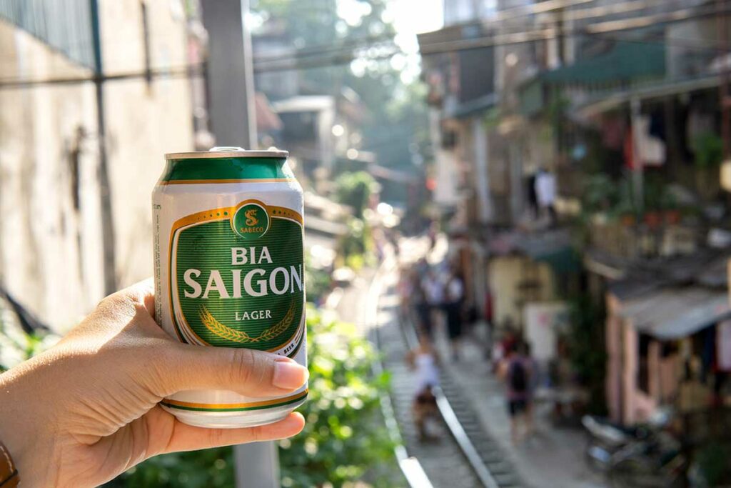 Hand met een blikje Bia Saigon bier bij Hanoi Train Street, een typisch moment van bier drinken in Vietnam op een levendige stedelijke locatie