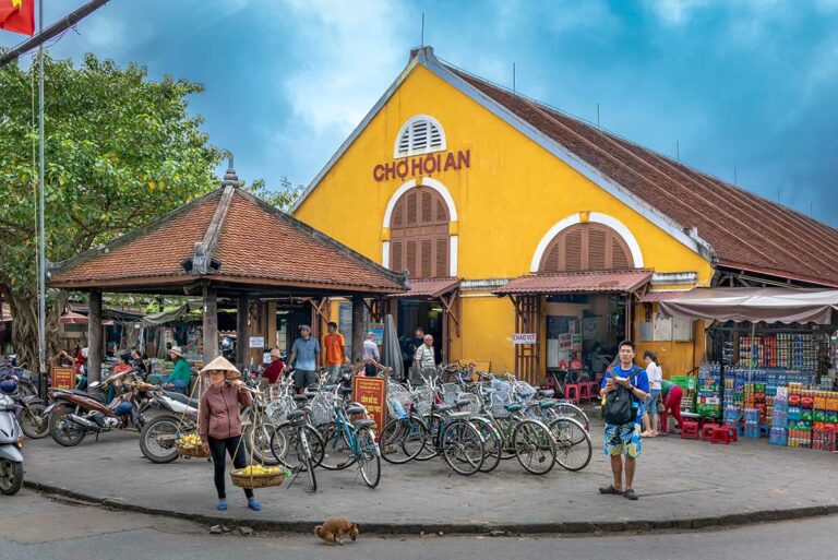 Buitenkant van de centrale markt van Hoi An, met geel koloniaal gebouw, fietsen, kraampjes en lokale handelaren in het stadscentrum