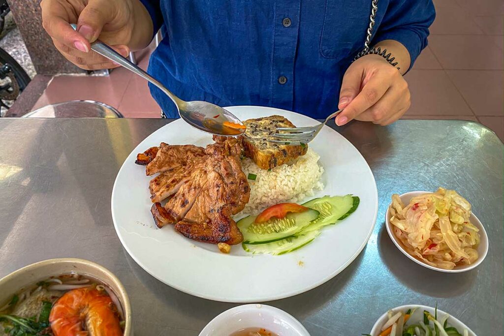 Cơm tấm, een Vietnamees gerecht met gebroken rijst, gegrild varkensvlees, gestoomde ei-vleesmengsel, verse komkommer en ingelegde groenten, geserveerd met vissaus