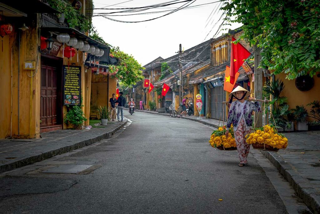 Vrouw met traditionele draagmand loopt door een rustige straat in de oude stad van Hoi An, een klassiek onderwerp voor straatfotografie in Vietnam.