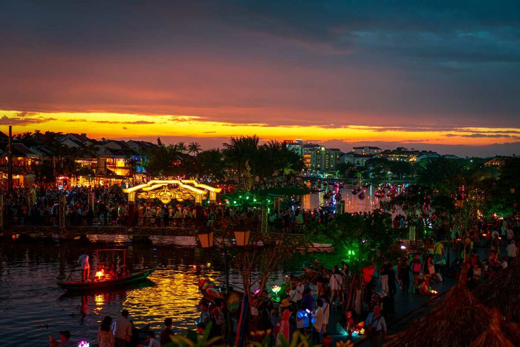 Grote menigte rond de An Hoi brug tijdens zonsondergang op het Hoi An Lantern Festival met boten en lampionnen op de rivier