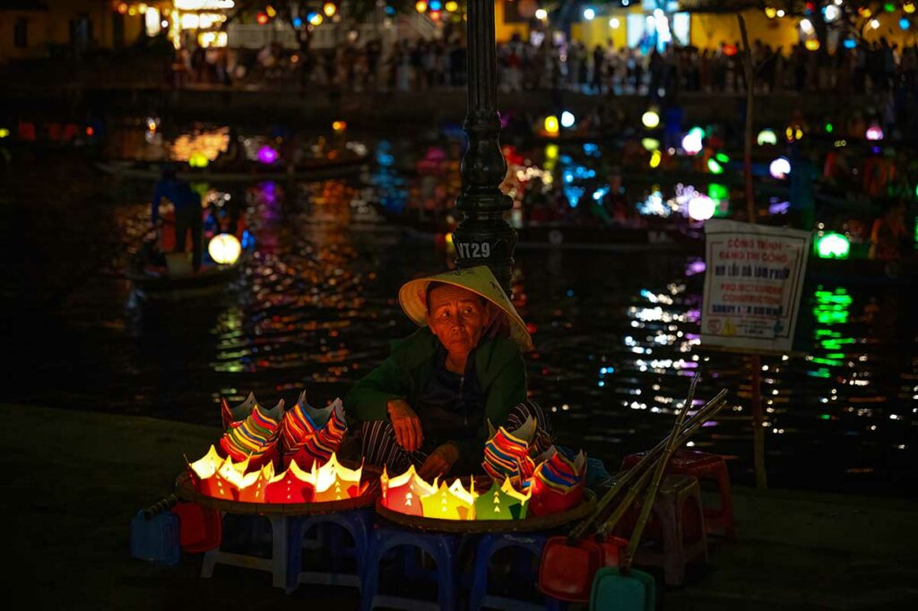 Oudere vrouw verkoopt papieren drijvende lampionnen langs de Hoai rivier tijdens het Hoi An Lantern Festival