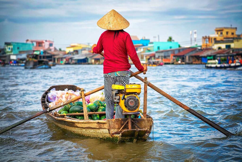 Vrouw staat op een kleine boot op de rivier in de Mekongdelta met goederen aan boord, een herkenbaar tafereel voor dagelijkse leven-fotografie in Vietnam.