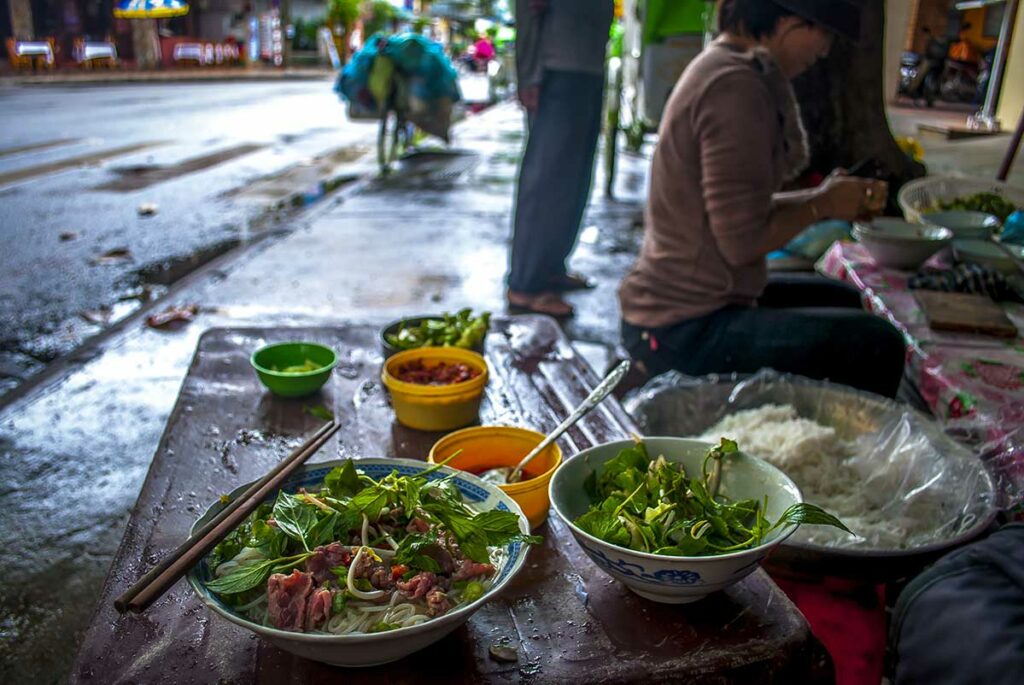 Kom phở met rijstnoedels, rundvlees en verse kruiden op een lage tafel langs de straat, een herkenbaar beeld van Vietnamees eten