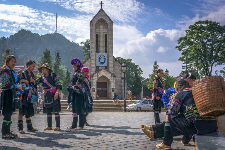 Hmong vrouwen in traditionele klederdracht op het plein voor de stenen kerk van Sapa, met de iconische katholieke kerk en berglandschap op de achtergrond