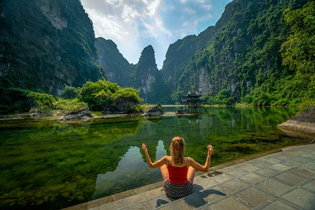 Reiziger zit aan de rand van een pad bij Trang An in Ninh Binh, omringd door groene kliffen en helder water, een rustig moment voor reisfotografie in Vietnam.