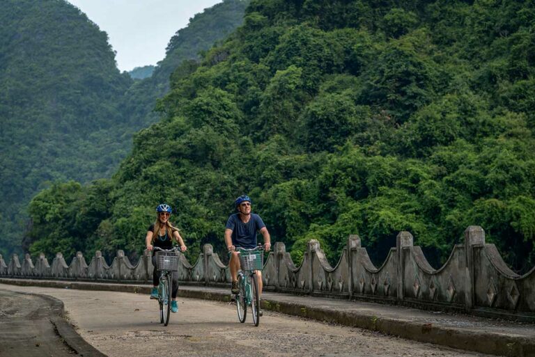 Twee reizigers fietsen door Viet Hai Village op Cat Ba Island, over een betonnen pad langs groene heuvels en water, een populaire activiteit in het landelijke deel van het eiland