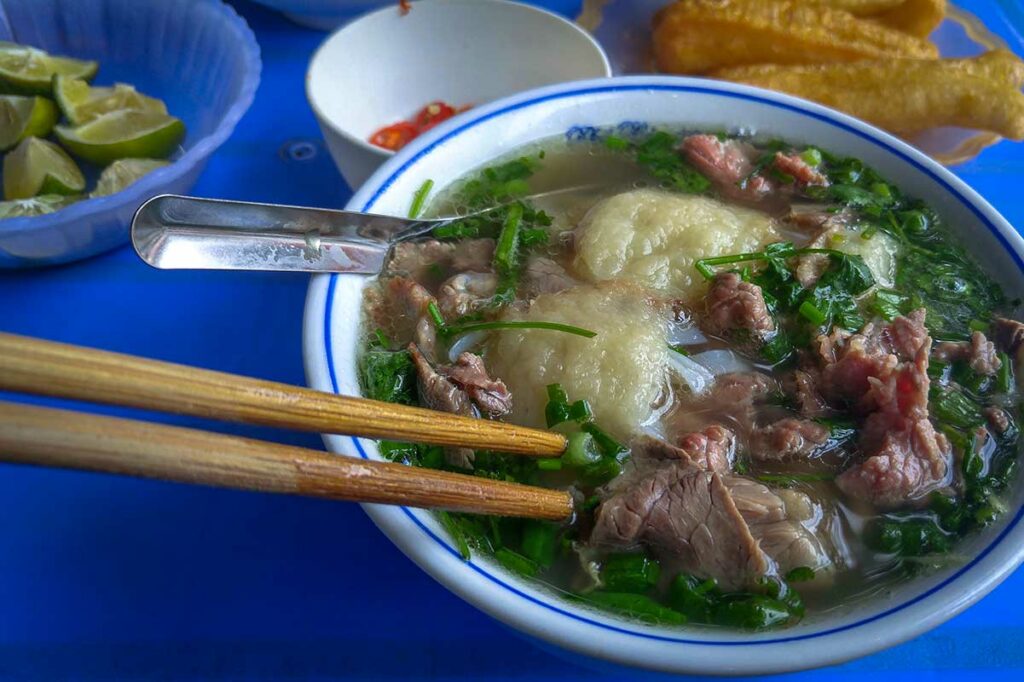 Kom Vietnamese phở met rundvlees, verse kruiden en rijstnoedels op een blauwe tafel, met citroen en chili ernaast om zelf toe te voegen, typisch street food in Vietnam