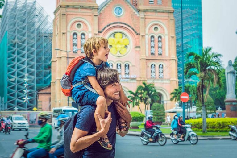 Vader met kind op schouders voor de Notre Dame kathedraal in Ho Chi Minh City met druk verkeer op de achtergrond