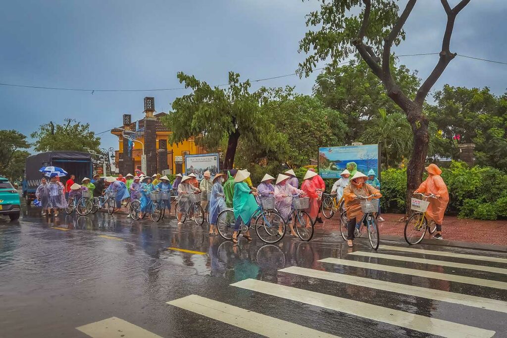 Toeristen fietsen door Hoi An in de regen met poncho’s tijdens het regenseizoen, typisch straatbeeld bij nat weer in Hoi An