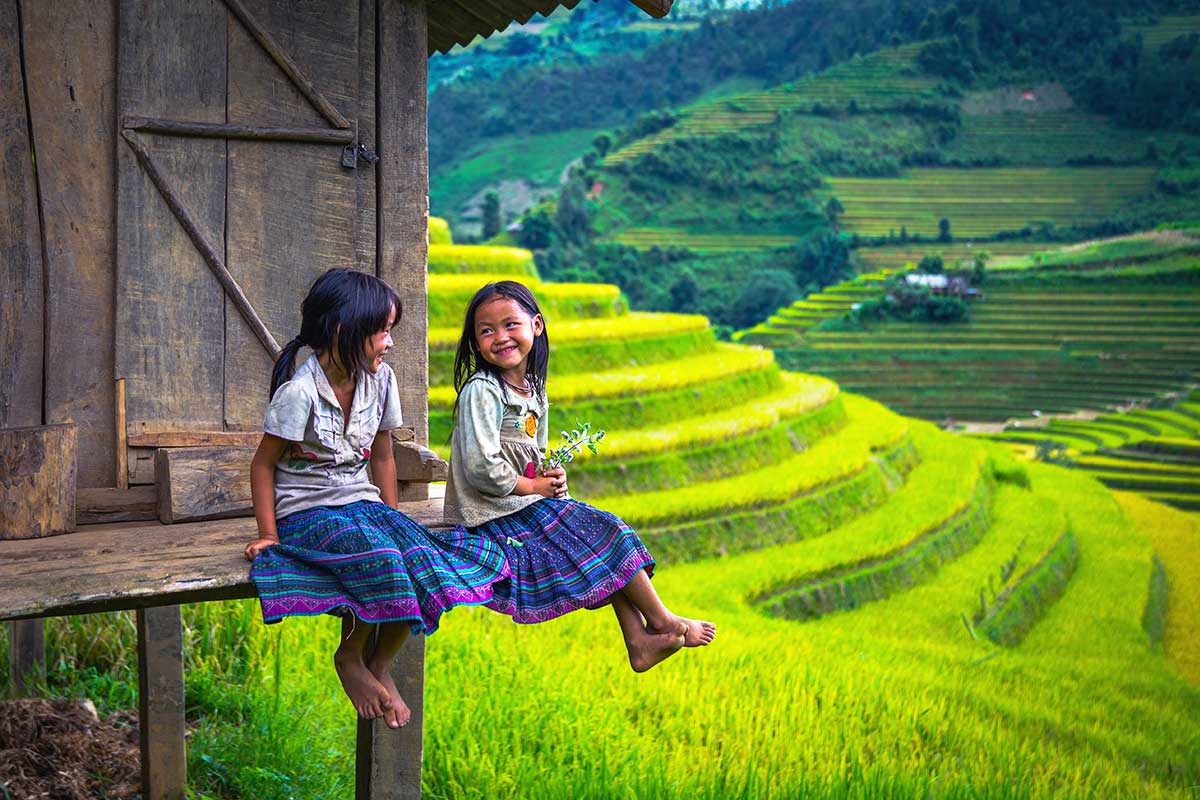 Twee kinderen zitten bij een huis op palen met op de achtergrond de terrasvormige rijstvelden van Sapa