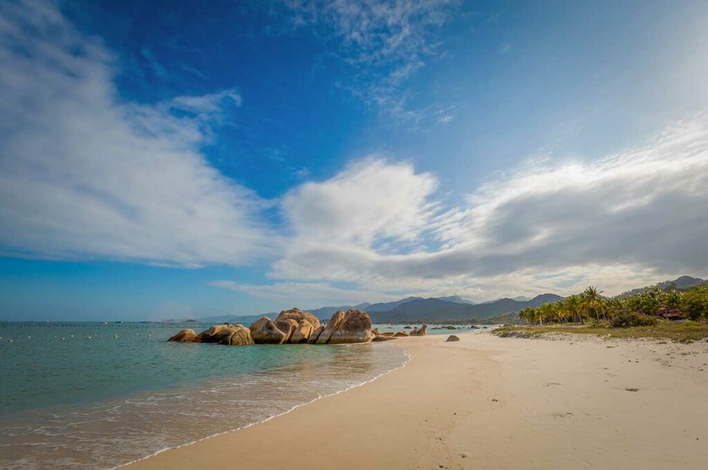 Rustig strand bij Cam Lam in Cam Ranh met wit zand, rotsformaties in zee en bergen op de achtergrond.
