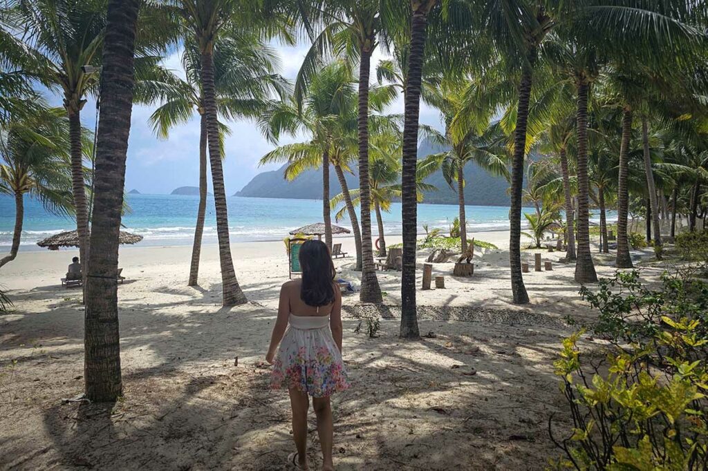 Vrouw loopt tussen palmbomen richting het strand van An Hai Beach in Con Dao met wit zand, ligbedden en helder blauw water.