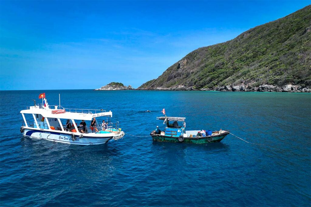 Duikboot en lokale vissersboot voor de kust van Con Dao, met helder blauw water en groene eilanden op de achtergrond, vertrekpunt voor duiken rond de stranden van Con Dao.