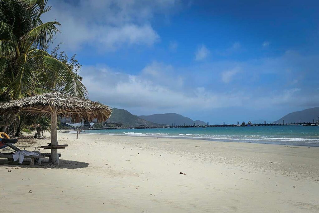 Rustig strand van An Hai Beach in Con Dao met wit zand, parasols van palmbladeren en uitzicht op de haven in de verte.