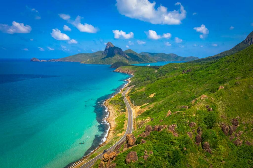 Kronkelende kustweg op Con Dao langs turquoise zee, groene heuvels en ruige rotsformaties, met uitzicht op de bergen in de verte.