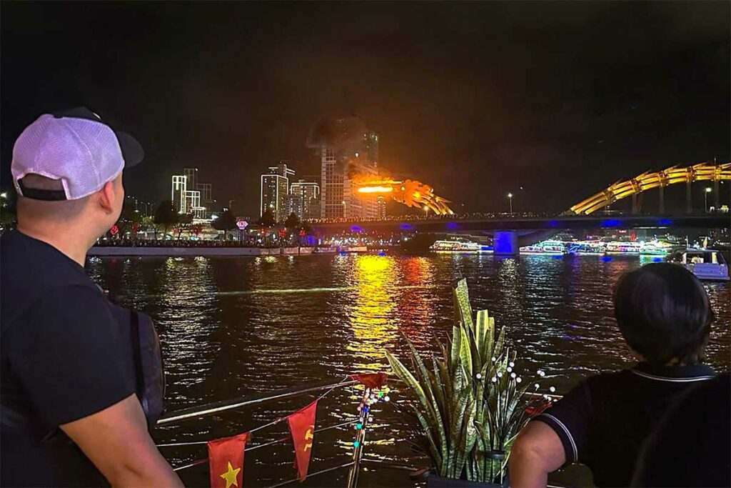 Uitzicht op de Dragon Bridge in Da Nang tijdens de vuurshow, gezien vanaf een boot op de Han River met reflecties van het vuur en stadslichten in het water.