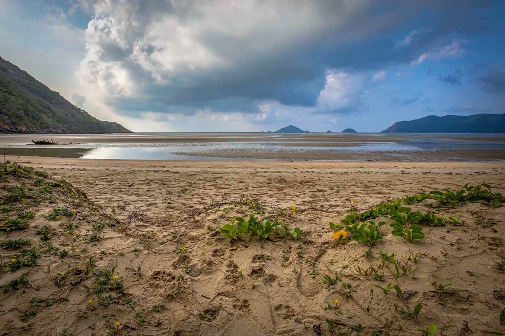 Rustig strand van Lo Voi Beach in Con Dao met nat zand bij eb, wolkenlucht en bergen langs de kustlijn.