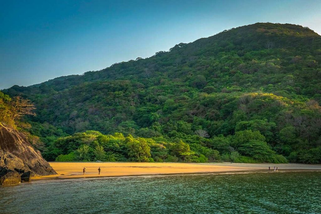 Rustig strand van Suoi Nong Beach in Con Dao met goudkleurig zand, enkele wandelaars en dichte groene jungle tegen de heuvels op de achtergrond.