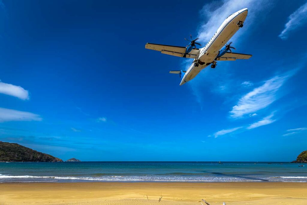 Passagiersvliegtuig dat over Dam Trau Beach in Con Dao vliegt vlak voor landing, gezien vanaf het strand met zee en bergen.