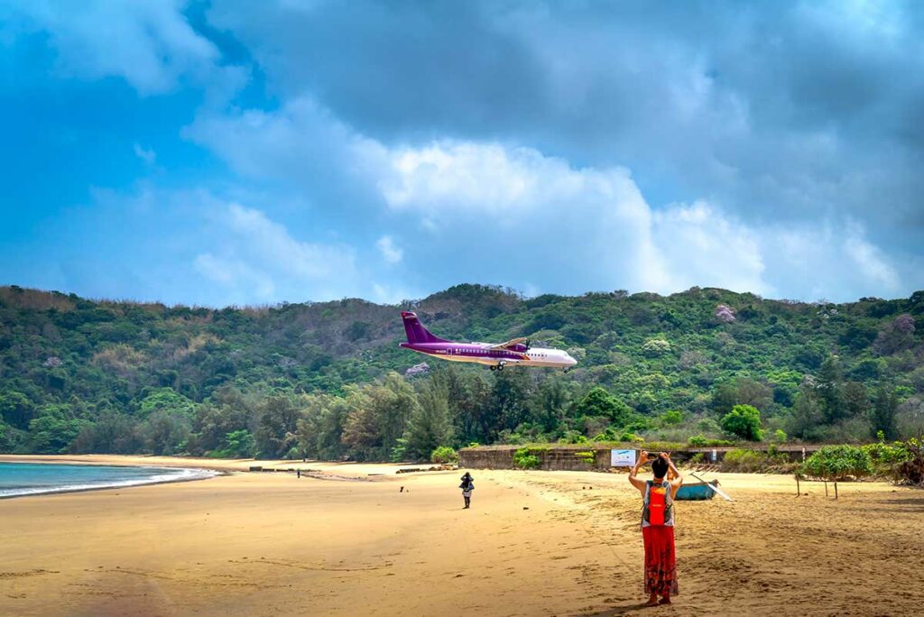Vliegtuig dat laag over Dam Trau Beach in Con Dao vliegt tijdens landing, met uitgestrekt zandstrand en bergen erachter.
