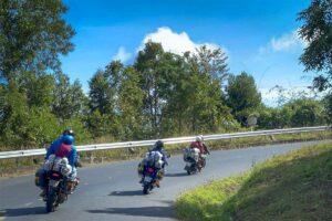 Drie motoren met reizigers achterop rijden in een bocht op een bergweg met bomen langs de kant bij Dalat — groepsritten zijn mogelijk bij een Easy Rider tour in de hooglanden rond Dalat