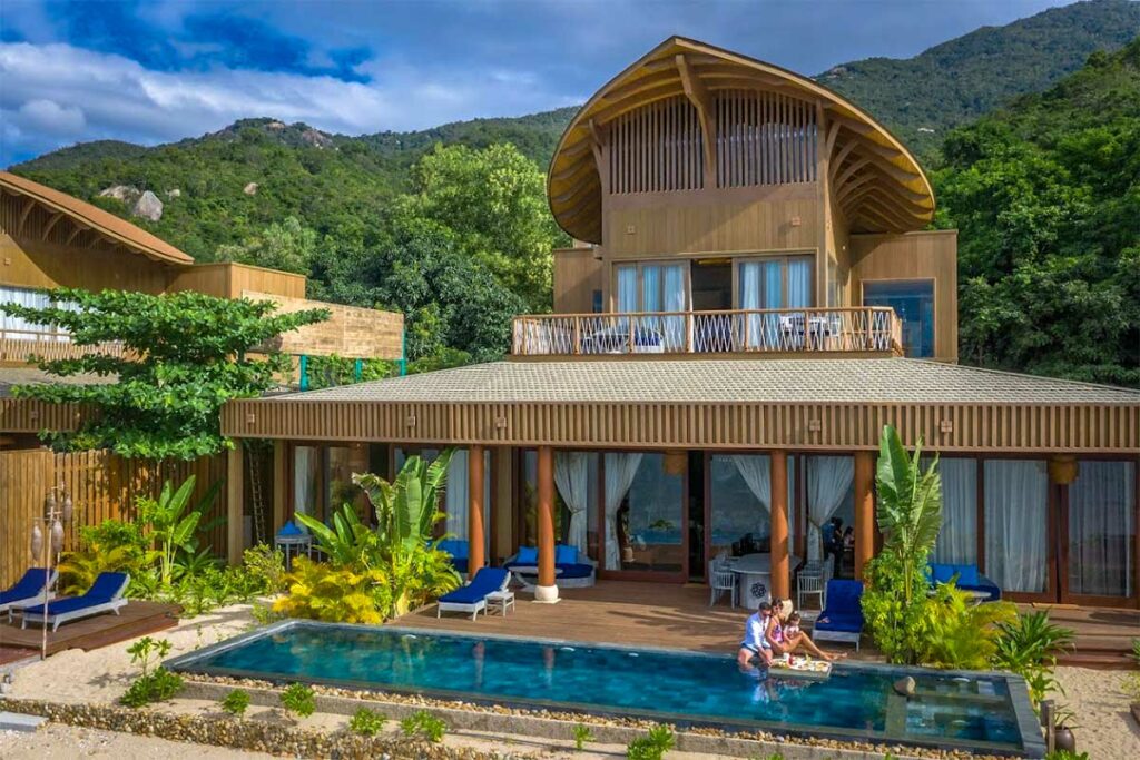 Villa van An Lam Retreats Ninh Van Bay met een gebogen houten dak, privézwembad en ligbedden op een terras, omgeven door tropische tuin en bergen — een van de luxueuze verblijven in de baai