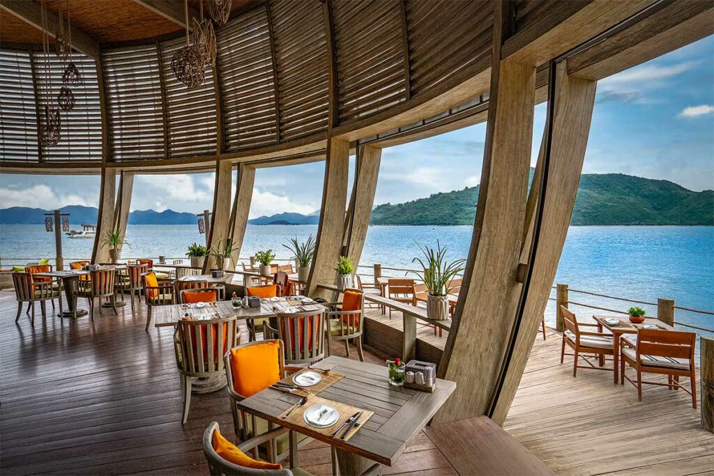 Open restaurant van An Lam Retreats Ninh Van Bay met houten stoelen met oranje kussens, panoramische ramen met uitzicht over de blauwe baai en eilanden in de verte