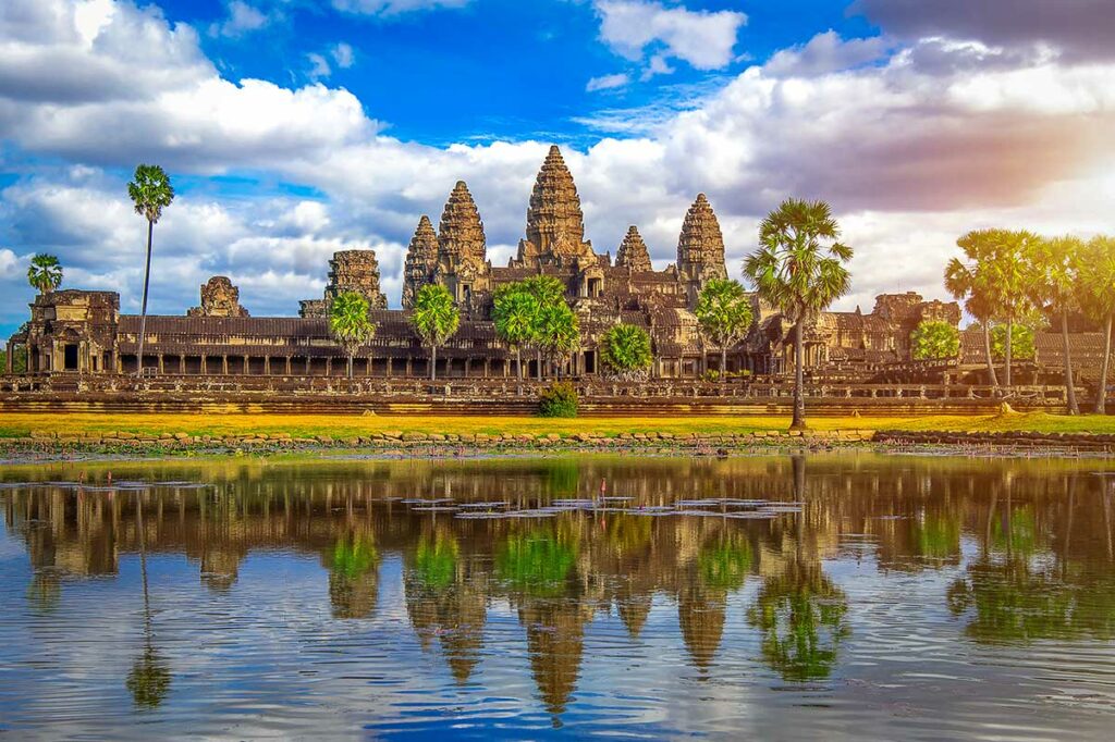Het tempelcomplex van Angkor Wat in Cambodja weerspiegeld in het water ervoor, omgeven door palmbomen onder een bewolkte lucht
