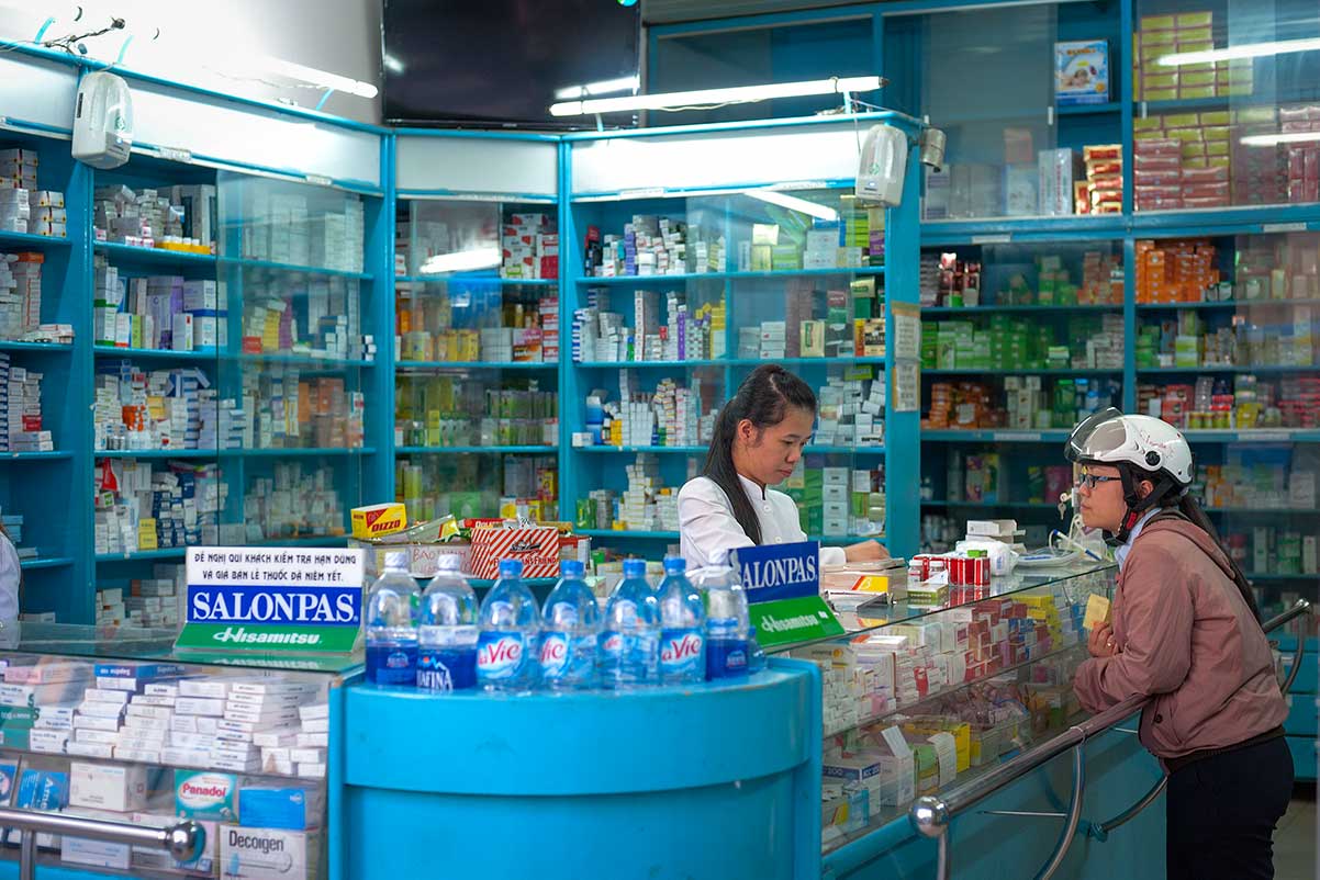 Lokale apotheek in Vietnam met een medewerker in witte jas achter de balie die een klant met motorhelm helpt, omringd door schappen vol medicijnen