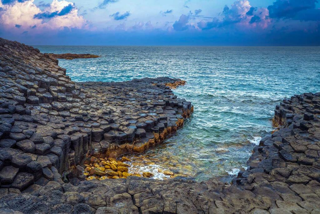 Zeshoekige basaltkolommen van het Da Dia rif in Phu Yen, gevormd door afgekoeld lavagesteente, met golven die tegen de rotsformaties slaan bij bewolkte zonsondergang — een geologisch bijzonder fenomeen aan de kust van Tuy Hoa