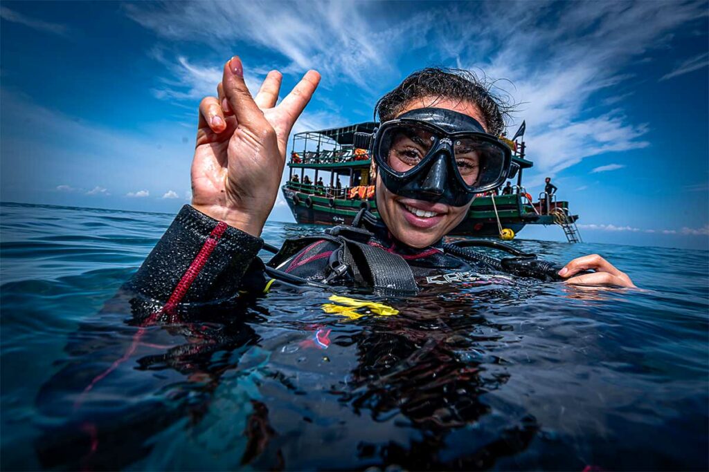 Duiker in wetsuit geeft het oké-teken aan de oppervlakte met een duikboot op de achtergrond bij de Cham eilanden — bootduiken is de standaard manier om de duikplekken rond Hoi An te bereiken