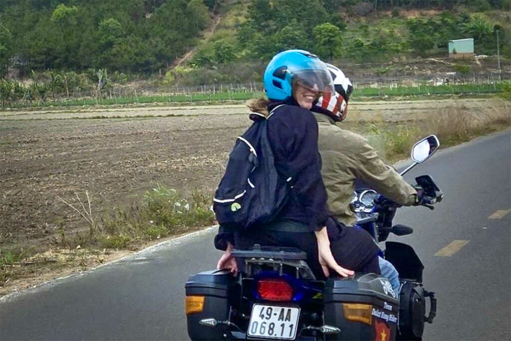 Reiziger kijkt lachend achterom terwijl ze achterop een Easy Rider-motor rijdt door het platteland bij Dalat, met akkers en heuvels op de achtergrond — het achterop rijden bij een lokale gids is de kern van een Easy Rider ervaring
