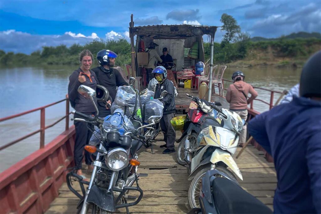 Reizigers en Easy Rider-gidsen met motoren op een kleine veerpont die een rivier oversteekt tijdens een meerdaagse Easy Rider tour vanuit Dalat — een typisch onderdeel van de minder bekende routes