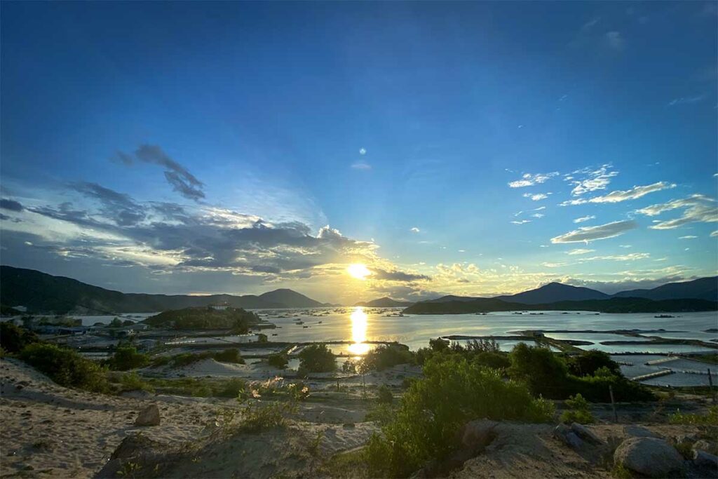 Zonsondergang boven de Van Phong Bay gezien vanaf het Dam Mon schiereiland, met viskweekinstallaties in het water, eilanden en bergen in de verte — een rustig en weinig bezocht uitkijkpunt