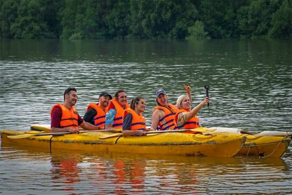 Groep reizigers in oranje zwemvesten kajakken in een grote gele kajak op de rivier bij Dan Xay in het Can Gio reservaat, met mangrovebos op de achtergrond — kajakken is een van de actievere manieren om het reservaat te verkennen