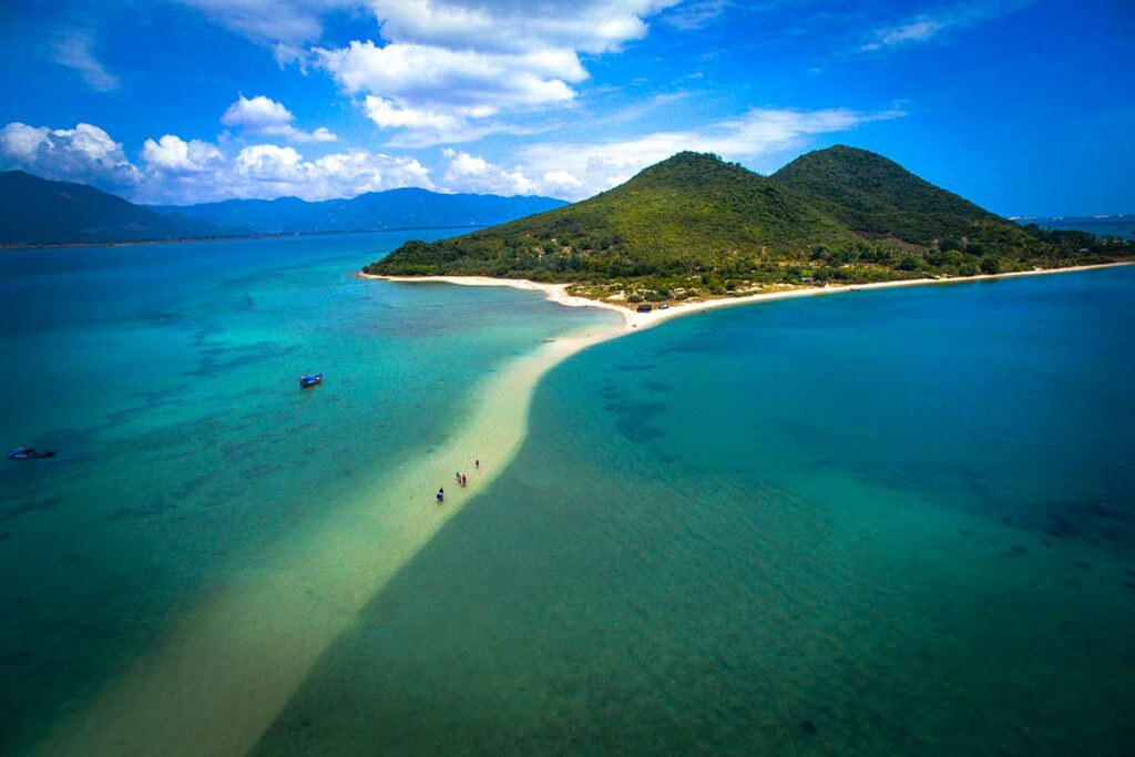 Luchtfoto van de smalle zandbank van Diep Son eiland in de Van Phong Bay, met bezoekers die over het ondiepe zand lopen tussen twee stukken turquoise water en een beboste heuvel op de achtergrond