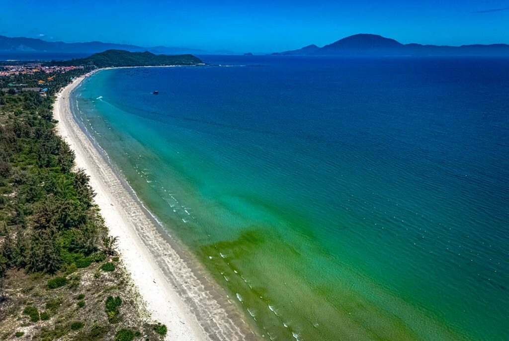 Luchtfoto van het lange witte strand van Doc Let Beach met helder groen en blauw water, een beboste kustlijn en bergen zichtbaar aan de horizon — een van de langste ongerepte stranden bij Nha Trang