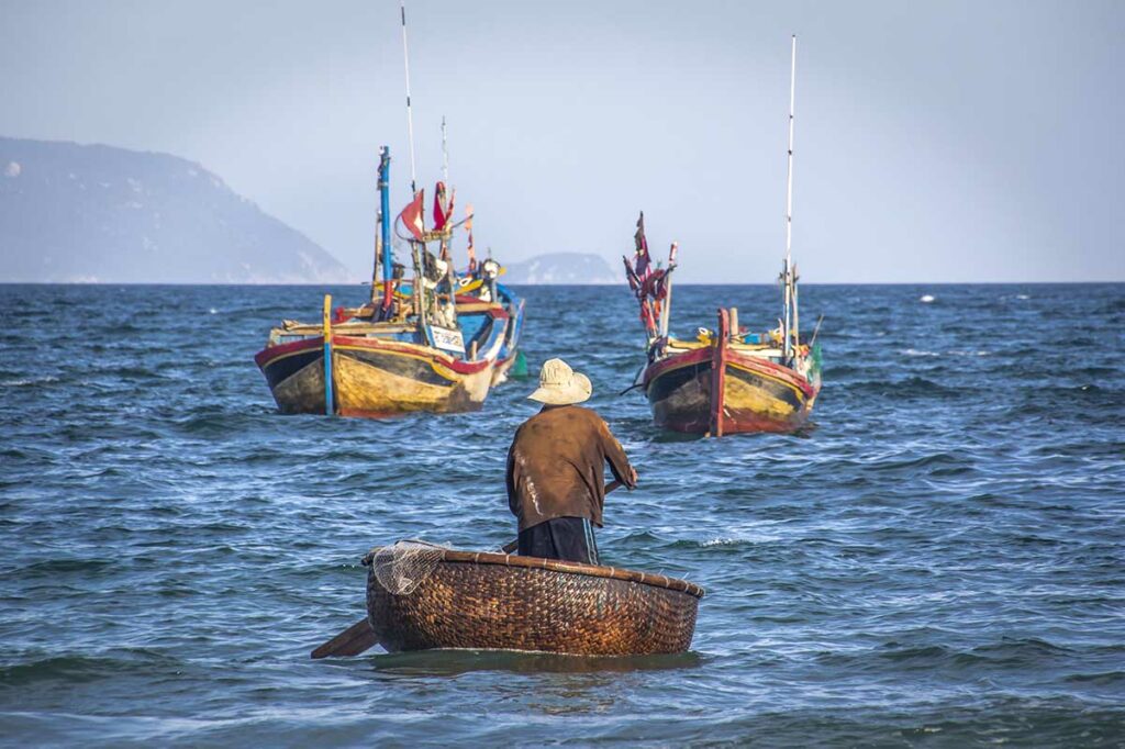 Vietnamese visser peddelt in een traditionele ronde coracle naar grotere houten vissersboten op zee bij Doc Let Beach — een authentiek beeld van het dagelijks leven langs deze kust